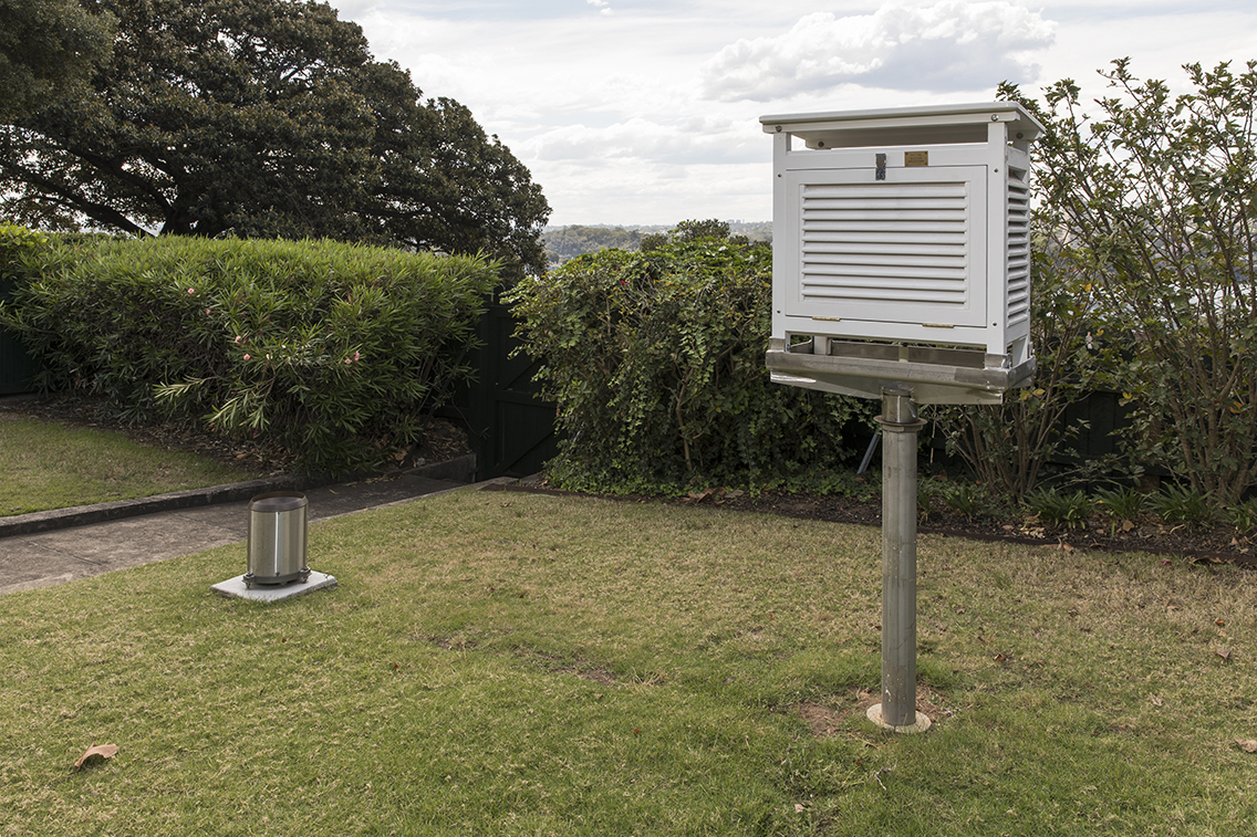 Lawn area in which is mounted a white box, approximately 40-50cm in size, on top of a metal post. The four sides of the box have louvres, fixed on an approximately 45 degree angle.